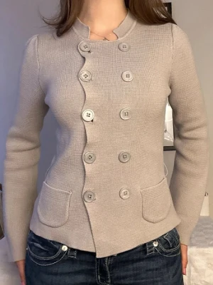 Ljusgrå elegant kofta🩶 - Stilren beige kofta med vågig knappslå och stora matchande knappar framtill. Koftan har två fickor fram, rund halsringning och är stickad i ett mjukt material. Perfekt för dig som gillar en clean och modern look.🩶