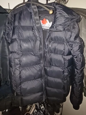 Svart dunjacka från Zavetti - En dragkedja är av    Snygg svart dunjacka från Zavetti med huva och dragkedja framtill. Jackan har en puffig design, diskret bröstficka med dragkedja och stor logga på ryggen. Perfekt för kalla vinterdagar och riktigt varm tack vare det tjocka materialet.