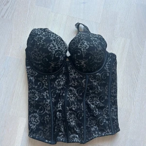 Svart spetskorsett med blommönster - Säljer en snygg svart korsett med silvrigt blommönster och vadderade kupor. Toppen har breda spetsband och markerade sömmar framtill för en cool siluett. Baksidan har justerbara band och hyskknäppning. Perfekt för dig som vill sticka ut med en edgy och feminin look.