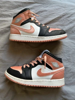 Air Jordan 1 Mid Peach/Black/White - Snygga Air Jordan 1 Mid sneakers i färgerna persika, svart och vitt. Skorna har klassisk Jordan-logga på sidan och tungan, samt persikofärgade snören och swoosh. Ovandelen är i textil och läder för en cool och sportig look.