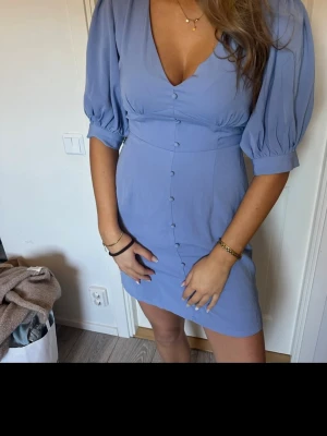 Ljusblå kort klänning med puffärm - Supersöt ljusblå klänning med puffärmar och v-ringning. Klänningen har knappar framtill och markerad midja som ger en feminin siluett. Perfekt för sommarens alla tillfällen och har en kort längd som känns både trendig och bekväm. Vet ej vart den är ifrån