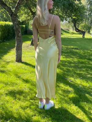 Ljusgul långklänning med smala band - Säljer en ljusgul långklänning i satinliknande material med smala axelband och öppen rygg med tunna banddetaljer. Använd endast 2 gånger