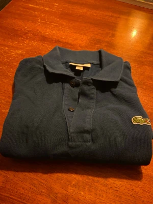 Blå slim fit piké från Lacoste - Säljer en klassisk blå pikétröja från Lacoste i slim fit-modell. Tröjan har krage, knappar framtill och den ikoniska krokodilloggan på bröstet. Perfekt för dig som gillar stilrena och sportiga plagg.