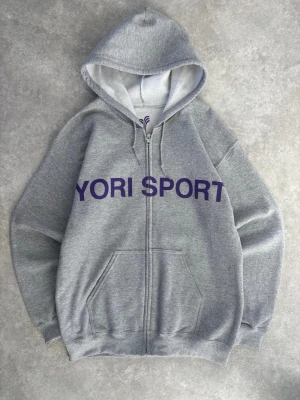 Grå hoodie YORI SPORT Crashoutboyz - Grå zip-hoodie från YORI SPORT med lila tryck framtill och stort tryck 'CRASHOUTBOYZ 77' på ryggen. Klassisk huva med dragsko och känguruficka. Perfekt för en avslappnad streetwear-look.