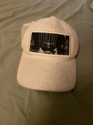 Vit truckerkeps med svartvit patch - Vit truckerkeps med mesh baktill och böjd skärm. Framtill sitter en svartvit patch med ett ikoniskt filmscenmotiv. Kepsen har justerbar storlek och är tillverkad i bomull och polyester.