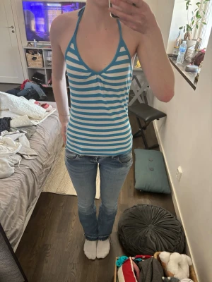 Blåvit randig halterneck topp - Säljer en blåvit randig halterneck linnetopp med smala axelband som knyts i nacken och djup urringning. Toppen är figurnära och har en öppen rygg, perfekt för varma dagar. Materialet känns mjukt och stretchigt, troligen bomull eller en bomullsblandning.