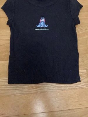 Svart ribbad t-shirt med Ior-tryck - Svart ribbad t-shirt med korta ärmar och ett tryck av Ior från Nalle Puh på bröstet samt texten 'Thanks for noticing me'. Perfekt för dig som gillar gulliga motiv och vill ha en enkel men söt t-shirt i garderoben.