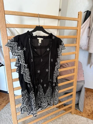 Svart blus med volang och vita broderade detaljer - Supersnygg svart blus med vita broderier och volangdetaljer längs ärmar och nederkant. Blusen har v-ringning med knytband och korta ärmar. Perfekt för dig som vill sticka ut med en unik topp!