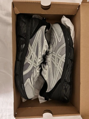 Asics X CP company grå/svarta sneakers - Snygga sneakers från Asics X CP company i grått och svart med coola mönster på ovandelen. Skorna har en chunky svart sula och meshmaterial som ger en sportig vibe. Perfekta för dig som gillar streetwear och vill ha något bekvämt och stilrent.