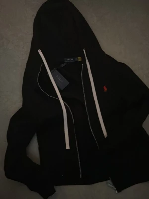 Svart Polo Ralph Lauren Hoddie  - Snygg svart hoodie från Polo Ralph Lauren med dragkedja, vita snören och den klassiska röda loggan broderad på bröstet. Tillverkad i mjukt material som känns skönt mot huden. Perfekt för en avslappnad och stilren look.
