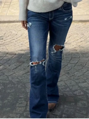 Blå bootcut jeans med slitningar - Snygga blå bootcut jeans med slitna detaljer och hål på knäna. Jeansen har vita kontrastsömmar och dekorativa broderier på bakfickorna. 