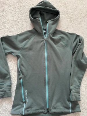 Grön fleecehoodie från Houdini - Mjuk och skön grön hoodie i fleece från Houdini med turkosa dragkedjor och två sidofickor. Hoodien har en stor huva och diskret logga på bröstet och huvan. Perfekt för lager-på-lager och chill dagar.