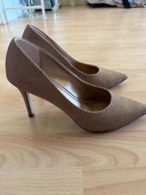Glittriga pumps från Catwalk - Säljer ett par glittriga pumps från Catwalk i en snygg guldig rosaktig nyans. Skorna har spetsig tå, smal klack och ett tyg med skimrande effekt som verkligen sticker ut. Perfekta för dig som vill ha något extra till outfiten.