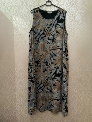 Svart ärmlös klänning med bladmotiv - Snygg ärmlös klänning med svart bas och mönster av beige och grå blad. Klänningen har rund halsringning och ett rakt, löst fall som ger en avslappnad siluett. Perfekt för dig som gillar naturinspirerade prints och vill ha något bekvämt men stilrent.