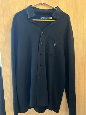 Svart skjorta från Polo Ralph Lauren - Klassisk svart skjorta från Polo Ralph Lauren med lång ärm och bröstficka. Skjortan har knappar framtill och den ikoniska loggan broderad på fickan. Perfekt till en stilren och avslappnad look.