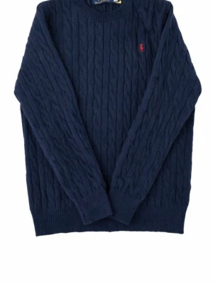 Mörkblå kabelstickad tröja Polo Ralph Lauren - Kabelstickad mörkblå tröja från Polo Ralph Lauren med rund halsringning och röd broderad logga på bröstet. Tröjan har långa ärmar och ribbade muddar vid ärmslut och nederkant. Klassisk och tidlös design i mjukt material.