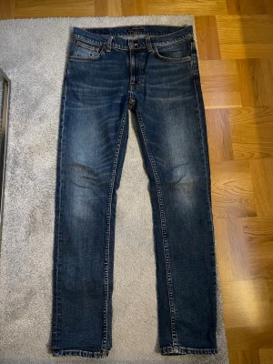 Mörkblå jeans från Nudie Jeans - Säljer ett par mörkblå jeans från Nudie Jeans med klassisk femficksmodell och orangea kontrastsömmar. Jeansen har en smal passform och är tillverkade i slitstark denim. Perfekta för dig som gillar en tidlös och stilren look.
