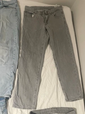 Grå raka jeans i klassisk modell - Säljer ett par grå jeans med rak passform och fem fickor. Jeansen har en klassisk look med normal midja och bälteshällor. Tillverkade i denim, perfekta för en avslappnad stil. Passar dig som gillar tidlös design och enkelhet.