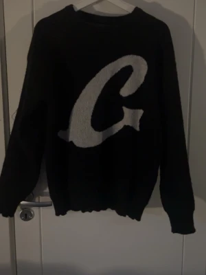 Svart stickad tröja med stor C-detalj - Stickad svart tröja med en stor vit bokstav C på framsidan. Tröjan har rund halsringning, långa ärmar och ribbade muddar vid ärmslut och nederkant. Perfekt för dig som gillar grafiska detaljer och enkel stil.