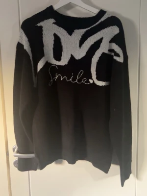Svart stickad tröja med vit text - Cool svart stickad tröja med vit grafisk text och ordet 'Smile' broderat framtill. Tröjan har rund halsringning och långa ärmar. Perfekt för dig som gillar streetstyle och vill ha något unikt i garderoben.