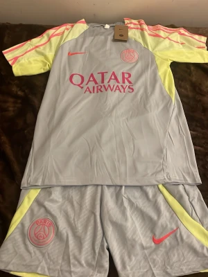 PSG träningsset Nike ljusgrå/rosa - Säljer ett PSG träningsset från Nike i ljusgrått med neon-gula och rosa detaljer. Tröjan har tryck med Qatar Airways och PSG-logga, samt rosa Nike swoosh. Matchande shorts med PSG-märke och rosa swoosh. Perfekt för fotbollsträning eller sport.
