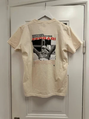 Michael Schumacher t-shirt - Snygg beige t-shirt med tryck av Michael Schumacher och Ferrari-logga på ryggen. T-shirten har korta ärmar och är tillverkad i mjuk bomull. Perfekt för dig som gillar motorsport och vill ha en unik och sportig stil.