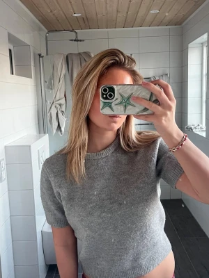 Grå stickad cropped tröja - Säljer en enkel och stilren grå stickad tröja med korta ärmar och cropped passform. Tröjan har rund halsringning och är gjord i ett mjukt material som passar perfekt till jeans eller kjol.