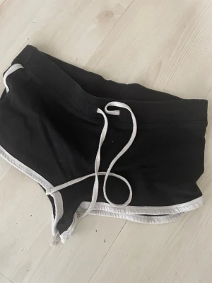 Svarta shorts med vit kant och snörning - Säljer ett par svarta shorts med vit kant och snörning i midjan. De har en sportig look och är perfekta för avslappnade dagar. Shortsen har en loose passform och är tillverkade i mjukt bomullsmaterial.   