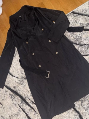 Svart trenchcoat  - Stilren svart trenchcoat från SHEIN i storlek petite XS.  Går ner till knäna på mig som är 157 cm. Skulle kunna passa barn också. Jackan har dubbelknäppning med silvriga knappar, klassisk krage och tillhörande bälte i midjan. Materialet är tunt och lätt, perfekt för lager på lager. Enkel och snygg design som passar till många outfits.