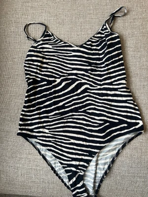 Zebramönstrad body från H&M - Snygg zebramönstrad body från H&M med tunna axelband och v-ringad hals. Toppen har svart och vit färg och är perfekt för dig som vill sticka ut med ett djurmönster. Materialet är mjukt och följsamt, vilket gör den skön att bära.