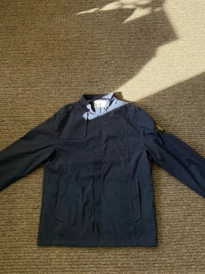 Mörkblå vindjacka från Stone Island - Snygg mörkblå vindjacka från Stone Island med klassisk patch på vänster ärm. Jackan har ståkrage, dragkedja framtill och två stora fickor. Materialet är tunt och vindtätt, perfekt för blåsiga dagar. Axelpartiet har dekorativa knappar.