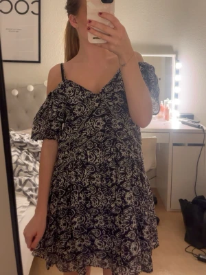 Vit kortklänning med marinblått mönster - Jag säljer den här korta klänning enmed svartvitt mönster och volangdetaljer. Klänningen har v-ringning, offshoulder och tunna axelband. Väldigt luftig och skön passform, passar perfekt för varma dagar där man bara vill ha något tunna på sig. Använd en gång. 