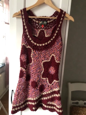 Bohemisk virkad klänning - Unik virkad klänning i vinröda, rosa och beige toner. Klänningen är ärmlös, kort och har stora blommiga mönster samt bohemiska detaljer. Perfekt för dig som gillar färg och retrostil. Materialet är stickad syntet och ullmix.