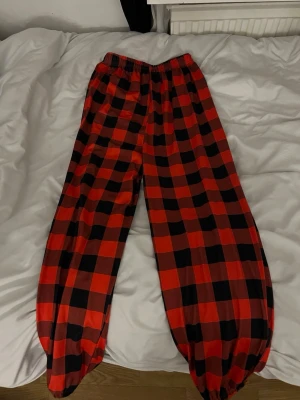 Rödrutiga pyjamasbyxor - Säljer ett par pyjamasbyxor med klassiskt röd- och svart rutigt mönster. Byxorna har resår i midjan och är gjorda i ett mjukt, glansigt material som känns skönt mot huden. Perfekta för mysiga hemmakvällar.