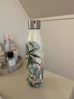 Vattenflaska med djungelmönster - Snygg vattenflaska i metall med tropiskt djungelmönster i grönt, rosa och vitt. Flaskan har ett silvrigt skruvlock och motiv av palmer, flamingos och blad. Perfekt att ta med till skolan eller gymmet.