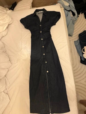 Mörkblå jeansklänning från Zara - Lång jeansklänning från Zara i mörkblå denim med korta ärmar och knappar hela vägen framtill. Klänningen har klassisk skjortkrage och markerad midja för en snygg siluett. Perfekt för dig som gillar en stilren och trendig look.