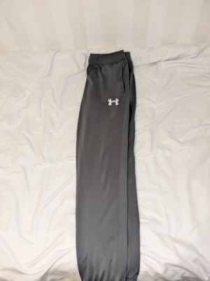 Grå mjukisbyxor från Under Armour - Säljer ett par grå mjukisbyxor från Under Armour med resår i midjan och vid bensluten. Byxorna har sidofickor och en vit logga på vänster lår. Perfekta för chill dagar eller träning.