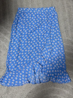 Blå blommig omlottkjol med volang - Supersöt blå omlottkjol med små vita och rosa blommor. Kjolen har volangkant nertill och är gjord i ett lätt, luftigt material som passar perfekt till varmare dagar. Passar även mindre.