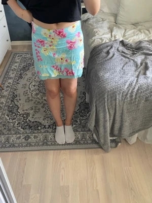 Blommig turkos kortkjol med volang - Supersöt kortkjol i turkos med blommigt mönster i rosa, gult och vitt. Kjolen har en volangkant nedtill. Materialet känns lätt och flowy, perfekt för sommaren.