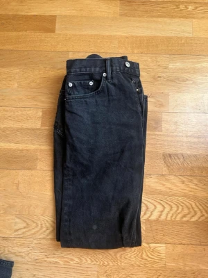 Svarta raka jeans från 157 - Säljer ett par klassiska svarta jeans från 157 med rak passform och fem fickor. Jeansen har silverfärgade nitar och knapp, samt bälteshällor. Tillverkade i kraftig denim som ger en tidlös look. Perfekta till en avslappnad streetstyle.
