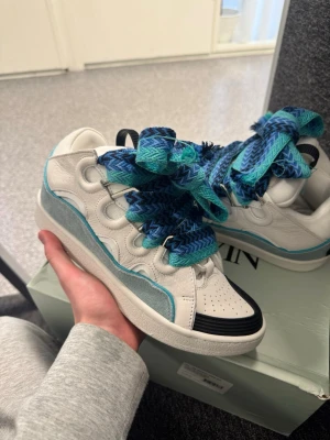 Lanvin Curbs - Unika sneakers från Lanvin i vitt läder med blå och turkosa detaljer samt breda, mönstrade skosnören i blått och grönt. Skon har en låg silhuett, svart tåparti och ljusblå mockadetalj längs sidan. Perfekt för dig som vill sticka ut med din stil.