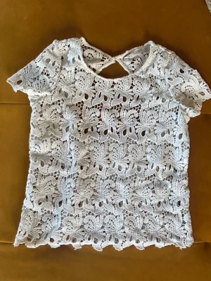 Vit spetsblus med öppen rygg  - Säljer en superfin vit blus i spets med korta ärmar och ett snyggt hålmönster. Toppen har öppen rygg och ett vackert blommigt mönster i spetsen. Perfekt att styla med jeans eller kjol.