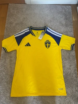 Sverige matchtröja gul Adidas - Gul matchtröja från Adidas med blå detaljer och Sveriges landslagsemblem på bröstet. Tröjan har korta ärmar, blåa axelpartier och vita ränder på axlarna. Tillverkad i lätt och ventilerande material, perfekt för fotboll eller träning.