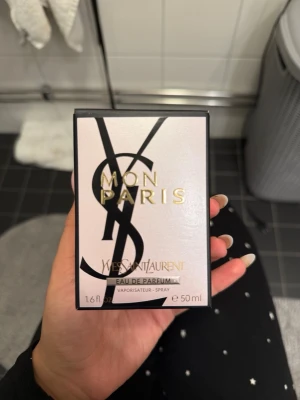 Yves Saint Laurent Mon Paris parfym - Mon Paris Eau de Parfum från Yves Saint Laurent i en elegant vit och svart kartong med guldtext. Parfymen är en spray och innehåller 50 ml. Förpackningen har en lyxig design med YSL-loggan i svart och detaljer i guld.