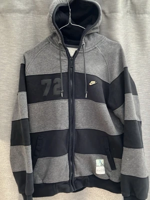 Grå och svart Nike hoodie med huva - Säljer en grå och svart hoodie från Nike med breda blockränder, huva med dragsko och hel dragkedja. Tryckt siffra 72 på bröstet och Nike-logga. Två fickor framtill och ribbade muddar. Perfekt för dig som gillar sportig och avslappnad stil.