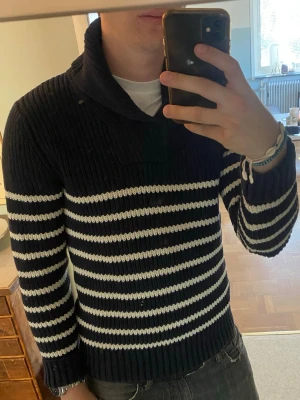 Marinblå stickad tröja med vita ränder - Säljer en marinblå stickad tröja med vita horisontella ränder. Tröjan har en hög krage och är ribbstickad för extra struktur. Perfekt för dig som gillar klassisk och stilren look. Materialet ser ut att vara tjockt och mjukt, troligen bomull eller ullblandning.