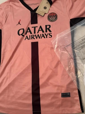 PSG x Jordan rosa matchtröja - Snygg rosa PSG matchtröja i samarbete med Jordan. Tröjan har svart vertikal rand, PSG-logga på bröstet och Qatar Airways som sponsortryck. Tillverkad i lätt och ventilerande Dri-FIT-material, perfekt för fotboll eller träning.
