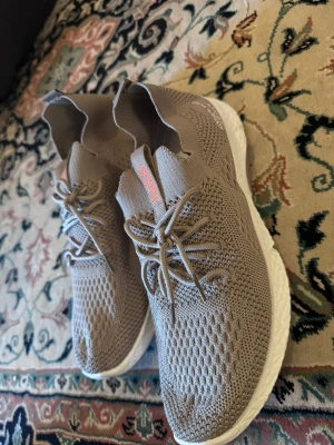 Beige stickade sneakers med vit sula - Säljer ett par beige sneakers från Sport med stickad ovandel och vita sulor. Skorna har snörning och en flexibel, strumpliknande passform. Perfekta för dig som gillar en modern och bekväm stil. Diskret logga på plösen i orange.