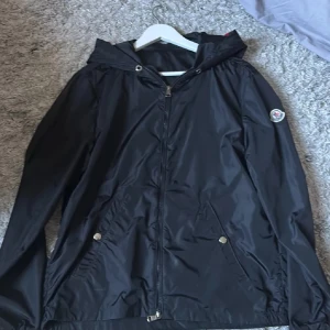 Moncler windbreaker  - Snygg svart vindjacka från Moncler med huva och dragkedja framtill. Jackan har två fickor med tryckknappar och Moncler-logga på ärmen. På huvan finns rödvita ränder som detalj. Tillverkad i ett glansigt, lätt material som står emot vind.