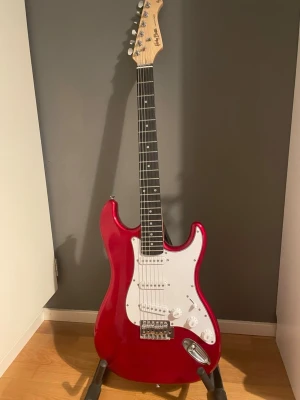 Röd elgitarr Harley Bentonio - Säljer en snygg röd elgitarr från Harley Benton med vit pickguard och klassisk Stratocaster-form. Gitarren har tre single coil-mikrofoner, svarta knappar och kromad stämskruv. Halsen är ljus trä och greppbrädan är mörk.  Ingår i gitarr högtalare / förstärkare och ett ställ för gitarren